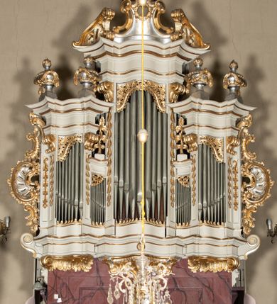 Zdjęcie nr 2: Szafa architektoniczna, pięcioosiowa, na falistym, profilowanym cokole, podtrzymywanym przez trzy ornamentalne wsporniki. Oś środkowa najwyższa, dwie skrajne niższe, ujęte pilastrami zakończonymi konsolami podtrzymującymi fragmenty falistego belkowania, z prześwitami na piszczałki w formie stojącego prostokąta. Osie środkowe najniższe, zakończone fragmentami niepełnego, falistego belkowania, z prześwitami na piszczałki w formie stojącego prostokąta. Oś środkowa zwieńczona ornamentalnym wolutowo-muszlowym szczytem z płaskorzeźbioną lirą oraz ornamentalnymi fragmentami przerwanego przyczółka. Na belkowaniu skrajnych osi ustawione cztery klasycystyczne wazony, a na belkowaniu osi środkowych cokoły z wicią roślinną. Trzony pilastrów dekorowane liściastymi zwisami. Prześwity na piszczałki w górnej części zdobione ornamentem roślinnym utworzonym z liści i kwiatów. Szafa flankowana rocaille'owymi uszami. Struktura polichromowana w kolorze białym, profile i ornamentyka złocone, detal srebrzony. 