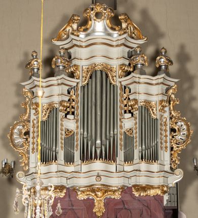 Zdjęcie nr 1: Szafa architektoniczna, pięcioosiowa, na falistym, profilowanym cokole, podtrzymywanym przez trzy ornamentalne wsporniki. Oś środkowa najwyższa, dwie skrajne niższe, ujęte pilastrami zakończonymi konsolami podtrzymującymi fragmenty falistego belkowania, z prześwitami na piszczałki w formie stojącego prostokąta. Osie środkowe najniższe, zakończone fragmentami niepełnego, falistego belkowania, z prześwitami na piszczałki w formie stojącego prostokąta. Oś środkowa zwieńczona ornamentalnym wolutowo-muszlowym szczytem z płaskorzeźbioną lirą oraz ornamentalnymi fragmentami przerwanego przyczółka. Na belkowaniu skrajnych osi ustawione cztery klasycystyczne wazony, a na belkowaniu osi środkowych cokoły z wicią roślinną. Trzony pilastrów dekorowane liściastymi zwisami. Prześwity na piszczałki w górnej części zdobione ornamentem roślinnym utworzonym z liści i kwiatów. Szafa flankowana rocaille&#039;owymi uszami. Struktura polichromowana w kolorze białym, profile i ornamentyka złocone, detal srebrzony. 