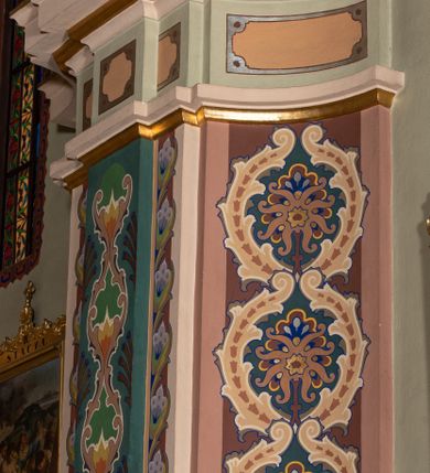 Zdjęcie nr 8: W prezbiterium w absydzie do dwóch trzecich wysokości ściany dekoracja roślinno-geometryczna z motywami kwiatowymi w kolorach zielono-turkusowym oraz złotym; powyżej dwa anioły na tle niebieskiego nieba z rozetami, całość zakończona zgeometryzowaną glorią promienistą uzupełnioną motywami kwiatowymi. 
Ściany boczne prezbiterium w kolorze jasnego różu; na ścianie tarczowej południowej medalion z kielichem i hostią w glorii promienistej. Na pilastrach, gurtach sklepiennych i w rozglifieniach okien symetryczna dekoracja roślinna. Na sklepieniu jasnopomarańczowe pole, w którego centrum znajduje się medalion z przedstawieniem Veraicon w glorii promienistej uzupełnionej motywami roślinnymi. 
Łuk tęczowy pokryty ornamentem roślinnym z motywami kłosów zbóż, na niebieskim tle, na środku łuku przedstawienie pelikana w gnieździe. Po wewnętrznej stronie łuku malatura w postaci fantazyjnego ornamentu roślinnego na beżowym tle. 
Ściany nawy gładkie, w kolorze zgaszonej, jasnej zieleni; z malowanymi zacheuszkami. W rozglifieniach okien symetryczna, stylizowana dekoracja arabeskowa na pomarańczowym i żółtym tle. Pilastry i łuki pokryte różnokolorowym, fantazyjnym ornamentem roślinnym. Sklepienia w każdym przęśle z wydzielonymi jasnożółtymi, owalnymi polami, w których centrum znajdują się niebieskie medaliony z przedstawieniem gorejącego serca lub monogramu IHS, w koronie cierniowej, w wieńcu z dekoracji roślinnej i w fioletowej glorii promienistej. W przyłuczach sklepień medaliony z przedstawieniami popiersi 12 apostołów, po czterech w każdym przęśle, ujętych żółtą wicią roślinną, na zielonym tle. Gurty sklepienne z motywem krzyży na żółto-pomarańczowym tle. 
Parapet chóru muzycznego i wspierające go arkady podzielone na malowane pola i płyciny wypełnione fantazyjnym ornamentem roślinnym. Na osi balustrady chóru przedstawienie liry. W podłuczu środkowej arkady pod chórem oko Opatrzności w wielobarwnej, stylizowanej glorii promienistej. 
Na ścianie zachodniej dwie półkoliście zamknięte nisze z przedstawieniami z lewej strony św. Cecylii grającej na organach, zaś z prawej króla Dawida grającego na harfie, na jasnoniebieskim tle. W rozglifieniach nisz stylizowana dekoracja geometryczno-roślinna. 
