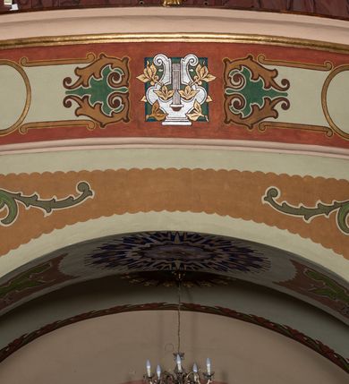 Zdjęcie nr 12: W prezbiterium w absydzie do dwóch trzecich wysokości ściany dekoracja roślinno-geometryczna z motywami kwiatowymi w kolorach zielono-turkusowym oraz złotym; powyżej dwa anioły na tle niebieskiego nieba z rozetami, całość zakończona zgeometryzowaną glorią promienistą uzupełnioną motywami kwiatowymi. 
Ściany boczne prezbiterium w kolorze jasnego różu; na ścianie tarczowej południowej medalion z kielichem i hostią w glorii promienistej. Na pilastrach, gurtach sklepiennych i w rozglifieniach okien symetryczna dekoracja roślinna. Na sklepieniu jasnopomarańczowe pole, w którego centrum znajduje się medalion z przedstawieniem Veraicon w glorii promienistej uzupełnionej motywami roślinnymi. 
Łuk tęczowy pokryty ornamentem roślinnym z motywami kłosów zbóż, na niebieskim tle, na środku łuku przedstawienie pelikana w gnieździe. Po wewnętrznej stronie łuku malatura w postaci fantazyjnego ornamentu roślinnego na beżowym tle. 
Ściany nawy gładkie, w kolorze zgaszonej, jasnej zieleni; z malowanymi zacheuszkami. W rozglifieniach okien symetryczna, stylizowana dekoracja arabeskowa na pomarańczowym i żółtym tle. Pilastry i łuki pokryte różnokolorowym, fantazyjnym ornamentem roślinnym. Sklepienia w każdym przęśle z wydzielonymi jasnożółtymi, owalnymi polami, w których centrum znajdują się niebieskie medaliony z przedstawieniem gorejącego serca lub monogramu IHS, w koronie cierniowej, w wieńcu z dekoracji roślinnej i w fioletowej glorii promienistej. W przyłuczach sklepień medaliony z przedstawieniami popiersi 12 apostołów, po czterech w każdym przęśle, ujętych żółtą wicią roślinną, na zielonym tle. Gurty sklepienne z motywem krzyży na żółto-pomarańczowym tle. 
Parapet chóru muzycznego i wspierające go arkady podzielone na malowane pola i płyciny wypełnione fantazyjnym ornamentem roślinnym. Na osi balustrady chóru przedstawienie liry. W podłuczu środkowej arkady pod chórem oko Opatrzności w wielobarwnej, stylizowanej glorii promienistej. 
Na ścianie zachodniej dwie półkoliście zamknięte nisze z przedstawieniami z lewej strony św. Cecylii grającej na organach, zaś z prawej króla Dawida grającego na harfie, na jasnoniebieskim tle. W rozglifieniach nisz stylizowana dekoracja geometryczno-roślinna. 
