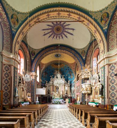 Zdjęcie nr 1: W prezbiterium w absydzie do dwóch trzecich wysokości ściany dekoracja roślinno-geometryczna z motywami kwiatowymi w kolorach zielono-turkusowym oraz złotym; powyżej dwa anioły na tle niebieskiego nieba z rozetami, całość zakończona zgeometryzowaną glorią promienistą uzupełnioną motywami kwiatowymi. 
Ściany boczne prezbiterium w kolorze jasnego różu; na ścianie tarczowej południowej medalion z kielichem i hostią w glorii promienistej. Na pilastrach, gurtach sklepiennych i w rozglifieniach okien symetryczna dekoracja roślinna. Na sklepieniu jasnopomarańczowe pole, w którego centrum znajduje się medalion z przedstawieniem Veraicon w glorii promienistej uzupełnionej motywami roślinnymi. 
Łuk tęczowy pokryty ornamentem roślinnym z motywami kłosów zbóż, na niebieskim tle, na środku łuku przedstawienie pelikana w gnieździe. Po wewnętrznej stronie łuku malatura w postaci fantazyjnego ornamentu roślinnego na beżowym tle. 
Ściany nawy gładkie, w kolorze zgaszonej, jasnej zieleni; z malowanymi zacheuszkami. W rozglifieniach okien symetryczna, stylizowana dekoracja arabeskowa na pomarańczowym i żółtym tle. Pilastry i łuki pokryte różnokolorowym, fantazyjnym ornamentem roślinnym. Sklepienia w każdym przęśle z wydzielonymi jasnożółtymi, owalnymi polami, w których centrum znajdują się niebieskie medaliony z przedstawieniem gorejącego serca lub monogramu IHS, w koronie cierniowej, w wieńcu z dekoracji roślinnej i w fioletowej glorii promienistej. W przyłuczach sklepień medaliony z przedstawieniami popiersi 12 apostołów, po czterech w każdym przęśle, ujętych żółtą wicią roślinną, na zielonym tle. Gurty sklepienne z motywem krzyży na żółto-pomarańczowym tle. 
Parapet chóru muzycznego i wspierające go arkady podzielone na malowane pola i płyciny wypełnione fantazyjnym ornamentem roślinnym. Na osi balustrady chóru przedstawienie liry. W podłuczu środkowej arkady pod chórem oko Opatrzności w wielobarwnej, stylizowanej glorii promienistej. 
Na ścianie zachodniej dwie półkoliście zamknięte nisze z przedstawieniami z lewej strony św. Cecylii grającej na organach, zaś z prawej króla Dawida grającego na harfie, na jasnoniebieskim tle. W rozglifieniach nisz stylizowana dekoracja geometryczno-roślinna. 
