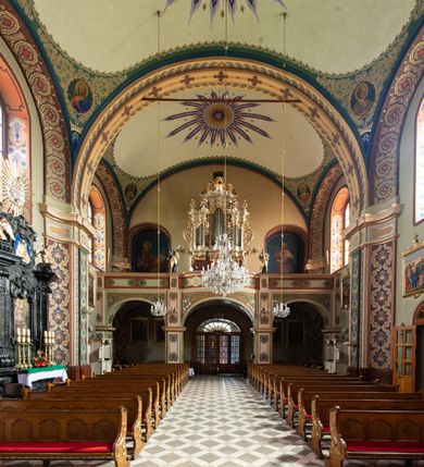 Zdjęcie nr 9: W prezbiterium w absydzie do dwóch trzecich wysokości ściany dekoracja roślinno-geometryczna z motywami kwiatowymi w kolorach zielono-turkusowym oraz złotym; powyżej dwa anioły na tle niebieskiego nieba z rozetami, całość zakończona zgeometryzowaną glorią promienistą uzupełnioną motywami kwiatowymi. 
Ściany boczne prezbiterium w kolorze jasnego różu; na ścianie tarczowej południowej medalion z kielichem i hostią w glorii promienistej. Na pilastrach, gurtach sklepiennych i w rozglifieniach okien symetryczna dekoracja roślinna. Na sklepieniu jasnopomarańczowe pole, w którego centrum znajduje się medalion z przedstawieniem Veraicon w glorii promienistej uzupełnionej motywami roślinnymi. 
Łuk tęczowy pokryty ornamentem roślinnym z motywami kłosów zbóż, na niebieskim tle, na środku łuku przedstawienie pelikana w gnieździe. Po wewnętrznej stronie łuku malatura w postaci fantazyjnego ornamentu roślinnego na beżowym tle. 
Ściany nawy gładkie, w kolorze zgaszonej, jasnej zieleni; z malowanymi zacheuszkami. W rozglifieniach okien symetryczna, stylizowana dekoracja arabeskowa na pomarańczowym i żółtym tle. Pilastry i łuki pokryte różnokolorowym, fantazyjnym ornamentem roślinnym. Sklepienia w każdym przęśle z wydzielonymi jasnożółtymi, owalnymi polami, w których centrum znajdują się niebieskie medaliony z przedstawieniem gorejącego serca lub monogramu IHS, w koronie cierniowej, w wieńcu z dekoracji roślinnej i w fioletowej glorii promienistej. W przyłuczach sklepień medaliony z przedstawieniami popiersi 12 apostołów, po czterech w każdym przęśle, ujętych żółtą wicią roślinną, na zielonym tle. Gurty sklepienne z motywem krzyży na żółto-pomarańczowym tle. 
Parapet chóru muzycznego i wspierające go arkady podzielone na malowane pola i płyciny wypełnione fantazyjnym ornamentem roślinnym. Na osi balustrady chóru przedstawienie liry. W podłuczu środkowej arkady pod chórem oko Opatrzności w wielobarwnej, stylizowanej glorii promienistej. 
Na ścianie zachodniej dwie półkoliście zamknięte nisze z przedstawieniami z lewej strony św. Cecylii grającej na organach, zaś z prawej króla Dawida grającego na harfie, na jasnoniebieskim tle. W rozglifieniach nisz stylizowana dekoracja geometryczno-roślinna. 
