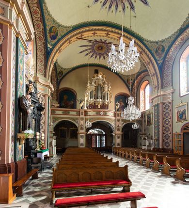 Zdjęcie nr 11: W prezbiterium w absydzie do dwóch trzecich wysokości ściany dekoracja roślinno-geometryczna z motywami kwiatowymi w kolorach zielono-turkusowym oraz złotym; powyżej dwa anioły na tle niebieskiego nieba z rozetami, całość zakończona zgeometryzowaną glorią promienistą uzupełnioną motywami kwiatowymi. 
Ściany boczne prezbiterium w kolorze jasnego różu; na ścianie tarczowej południowej medalion z kielichem i hostią w glorii promienistej. Na pilastrach, gurtach sklepiennych i w rozglifieniach okien symetryczna dekoracja roślinna. Na sklepieniu jasnopomarańczowe pole, w którego centrum znajduje się medalion z przedstawieniem Veraicon w glorii promienistej uzupełnionej motywami roślinnymi. 
Łuk tęczowy pokryty ornamentem roślinnym z motywami kłosów zbóż, na niebieskim tle, na środku łuku przedstawienie pelikana w gnieździe. Po wewnętrznej stronie łuku malatura w postaci fantazyjnego ornamentu roślinnego na beżowym tle. 
Ściany nawy gładkie, w kolorze zgaszonej, jasnej zieleni; z malowanymi zacheuszkami. W rozglifieniach okien symetryczna, stylizowana dekoracja arabeskowa na pomarańczowym i żółtym tle. Pilastry i łuki pokryte różnokolorowym, fantazyjnym ornamentem roślinnym. Sklepienia w każdym przęśle z wydzielonymi jasnożółtymi, owalnymi polami, w których centrum znajdują się niebieskie medaliony z przedstawieniem gorejącego serca lub monogramu IHS, w koronie cierniowej, w wieńcu z dekoracji roślinnej i w fioletowej glorii promienistej. W przyłuczach sklepień medaliony z przedstawieniami popiersi 12 apostołów, po czterech w każdym przęśle, ujętych żółtą wicią roślinną, na zielonym tle. Gurty sklepienne z motywem krzyży na żółto-pomarańczowym tle. 
Parapet chóru muzycznego i wspierające go arkady podzielone na malowane pola i płyciny wypełnione fantazyjnym ornamentem roślinnym. Na osi balustrady chóru przedstawienie liry. W podłuczu środkowej arkady pod chórem oko Opatrzności w wielobarwnej, stylizowanej glorii promienistej. 
Na ścianie zachodniej dwie półkoliście zamknięte nisze z przedstawieniami z lewej strony św. Cecylii grającej na organach, zaś z prawej króla Dawida grającego na harfie, na jasnoniebieskim tle. W rozglifieniach nisz stylizowana dekoracja geometryczno-roślinna. 
