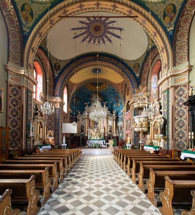 Zdjęcie nr 2: W prezbiterium w absydzie do dwóch trzecich wysokości ściany dekoracja roślinno-geometryczna z motywami kwiatowymi w kolorach zielono-turkusowym oraz złotym; powyżej dwa anioły na tle niebieskiego nieba z rozetami, całość zakończona zgeometryzowaną glorią promienistą uzupełnioną motywami kwiatowymi. 
Ściany boczne prezbiterium w kolorze jasnego różu; na ścianie tarczowej południowej medalion z kielichem i hostią w glorii promienistej. Na pilastrach, gurtach sklepiennych i w rozglifieniach okien symetryczna dekoracja roślinna. Na sklepieniu jasnopomarańczowe pole, w którego centrum znajduje się medalion z przedstawieniem Veraicon w glorii promienistej uzupełnionej motywami roślinnymi. 
Łuk tęczowy pokryty ornamentem roślinnym z motywami kłosów zbóż, na niebieskim tle, na środku łuku przedstawienie pelikana w gnieździe. Po wewnętrznej stronie łuku malatura w postaci fantazyjnego ornamentu roślinnego na beżowym tle. 
Ściany nawy gładkie, w kolorze zgaszonej, jasnej zieleni; z malowanymi zacheuszkami. W rozglifieniach okien symetryczna, stylizowana dekoracja arabeskowa na pomarańczowym i żółtym tle. Pilastry i łuki pokryte różnokolorowym, fantazyjnym ornamentem roślinnym. Sklepienia w każdym przęśle z wydzielonymi jasnożółtymi, owalnymi polami, w których centrum znajdują się niebieskie medaliony z przedstawieniem gorejącego serca lub monogramu IHS, w koronie cierniowej, w wieńcu z dekoracji roślinnej i w fioletowej glorii promienistej. W przyłuczach sklepień medaliony z przedstawieniami popiersi 12 apostołów, po czterech w każdym przęśle, ujętych żółtą wicią roślinną, na zielonym tle. Gurty sklepienne z motywem krzyży na żółto-pomarańczowym tle. 
Parapet chóru muzycznego i wspierające go arkady podzielone na malowane pola i płyciny wypełnione fantazyjnym ornamentem roślinnym. Na osi balustrady chóru przedstawienie liry. W podłuczu środkowej arkady pod chórem oko Opatrzności w wielobarwnej, stylizowanej glorii promienistej. 
Na ścianie zachodniej dwie półkoliście zamknięte nisze z przedstawieniami z lewej strony św. Cecylii grającej na organach, zaś z prawej króla Dawida grającego na harfie, na jasnoniebieskim tle. W rozglifieniach nisz stylizowana dekoracja geometryczno-roślinna. 
