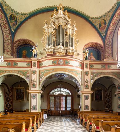 Zdjęcie nr 10: W prezbiterium w absydzie do dwóch trzecich wysokości ściany dekoracja roślinno-geometryczna z motywami kwiatowymi w kolorach zielono-turkusowym oraz złotym; powyżej dwa anioły na tle niebieskiego nieba z rozetami, całość zakończona zgeometryzowaną glorią promienistą uzupełnioną motywami kwiatowymi. 
Ściany boczne prezbiterium w kolorze jasnego różu; na ścianie tarczowej południowej medalion z kielichem i hostią w glorii promienistej. Na pilastrach, gurtach sklepiennych i w rozglifieniach okien symetryczna dekoracja roślinna. Na sklepieniu jasnopomarańczowe pole, w którego centrum znajduje się medalion z przedstawieniem Veraicon w glorii promienistej uzupełnionej motywami roślinnymi. 
Łuk tęczowy pokryty ornamentem roślinnym z motywami kłosów zbóż, na niebieskim tle, na środku łuku przedstawienie pelikana w gnieździe. Po wewnętrznej stronie łuku malatura w postaci fantazyjnego ornamentu roślinnego na beżowym tle. 
Ściany nawy gładkie, w kolorze zgaszonej, jasnej zieleni; z malowanymi zacheuszkami. W rozglifieniach okien symetryczna, stylizowana dekoracja arabeskowa na pomarańczowym i żółtym tle. Pilastry i łuki pokryte różnokolorowym, fantazyjnym ornamentem roślinnym. Sklepienia w każdym przęśle z wydzielonymi jasnożółtymi, owalnymi polami, w których centrum znajdują się niebieskie medaliony z przedstawieniem gorejącego serca lub monogramu IHS, w koronie cierniowej, w wieńcu z dekoracji roślinnej i w fioletowej glorii promienistej. W przyłuczach sklepień medaliony z przedstawieniami popiersi 12 apostołów, po czterech w każdym przęśle, ujętych żółtą wicią roślinną, na zielonym tle. Gurty sklepienne z motywem krzyży na żółto-pomarańczowym tle. 
Parapet chóru muzycznego i wspierające go arkady podzielone na malowane pola i płyciny wypełnione fantazyjnym ornamentem roślinnym. Na osi balustrady chóru przedstawienie liry. W podłuczu środkowej arkady pod chórem oko Opatrzności w wielobarwnej, stylizowanej glorii promienistej. 
Na ścianie zachodniej dwie półkoliście zamknięte nisze z przedstawieniami z lewej strony św. Cecylii grającej na organach, zaś z prawej króla Dawida grającego na harfie, na jasnoniebieskim tle. W rozglifieniach nisz stylizowana dekoracja geometryczno-roślinna. 
