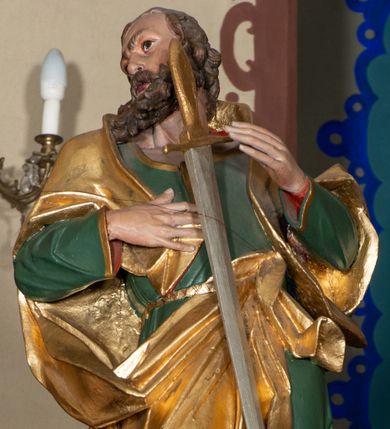 Zdjęcie nr 1: Rzeźba pełna, ustawiona na niskiej podstawie. Święty zwrócony frontalnie, z głową skierowaną w prawo, w kontrapoście, z silnie wypchniętym w bok lewym biodrem. Prawą rękę kładzie na piersi, lewą ugiętą unosi. Na tle piersi zamontowany miecz, skierowany ostrzem w dół. Twarz ma szczupłą, o dużych oczach, garbatym nosie i pełnych, rozchylonych ustach; okoloną gęstą brodą oraz krótkimi, kręconymi włosami. Ubrany jest w zieloną tunikę o złoconej lamówce, przepasaną w talii, odsłaniającą prawą nogę oraz złocony płaszcz narzucony na ramiona i zawieszony przy pasie z przodu postaci. Na stopach ma złocone buty z wysokimi, wywiniętymi cholewkami. Polichromia ciała naturalistyczna. 
