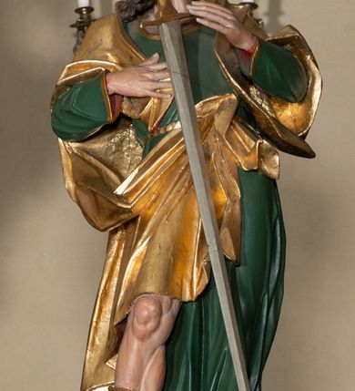 Zdjęcie nr 2: Rzeźba pełna, ustawiona na niskiej podstawie. Święty zwrócony frontalnie, z głową skierowaną w prawo, w kontrapoście, z silnie wypchniętym w bok lewym biodrem. Prawą rękę kładzie na piersi, lewą ugiętą unosi. Na tle piersi zamontowany miecz, skierowany ostrzem w dół. Twarz ma szczupłą, o dużych oczach, garbatym nosie i pełnych, rozchylonych ustach; okoloną gęstą brodą oraz krótkimi, kręconymi włosami. Ubrany jest w zieloną tunikę o złoconej lamówce, przepasaną w talii, odsłaniającą prawą nogę oraz złocony płaszcz narzucony na ramiona i zawieszony przy pasie z przodu postaci. Na stopach ma złocone buty z wysokimi, wywiniętymi cholewkami. Polichromia ciała naturalistyczna. 
