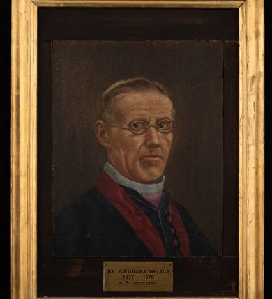 Zdjęcie nr 2: Obraz uzyskał kształt stojącego prostokąta, który został powiększony przez dodanie po bokach wstawek, umożliwiających jego oprawę w profilowaną, złoconą ramę, wiekszą niż sam portret. W centrum kompozycji ukazano na ciemnym tle w popiersiu duchownego w trzech czwartych zwróconego w lewo. Jest to starszy mężczyzna w okrągłych okularach w złoconych oprawkach, o pociągłej twarzy z wysokim czołem z widocznymi na twarzy zmarszczkami i siwiejącymi włosami zaczesanymi do tyłu odsłaniającymi prawe ucho. Ma wydatny nos i wargi. Ubrany jest w ciemnogranatową czamarę zapinaną rzędem filetowych guziczków widocznych z przodu. Spod niej widoczna jest biała koloratka w fioletowym pektorale. Na szyi widoczny jest fragment czerwonej wstęgi od dystynktorium. Pod portretem umieszczono tabliczkę z napisem "Ks(iądz) ANDRZEJ BYLICA / 1877-1878 / w Brzeszczach". 
