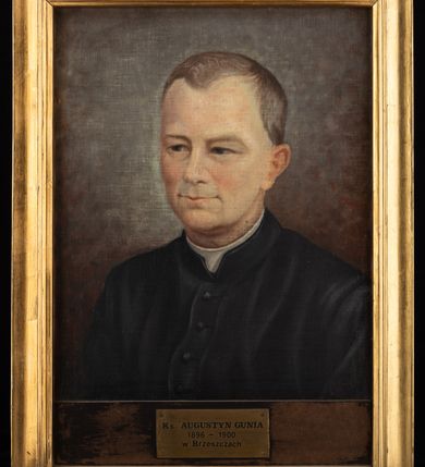 Zdjęcie nr 2: Portret uzyskał kształt stojącego prostokąta ujętego w złoconą, profilowaną ramę. Na ciemnym tle przedstawiono w popiersiu duchownego ukazanego w trzech czwartych w prawo. Jest to dojrzały mężczyzna z pociągłą twarzą z wysokim czołem z widocznymi zakolami siwiejących, krótkich włosów zaczesanych w niewielką grzywkę i odsłaniających lewe ucho. Jego twarz charakteryzują jasna karnacja, wydatny nos, wąskie usta i migdałowe ciemne oczy. Ubrany jest w czarną sutannę z białą koloratką pod szyją. Pod portretem umieszczono tabliczkę z napisem "Ks(iądz) AUGUSTYN GUNIA / 1896-1900 / w Brzeszczach". 