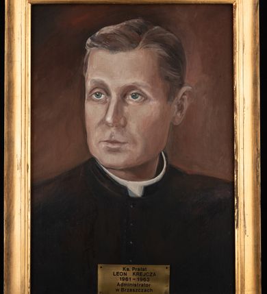 Zdjęcie nr 2: Portret uzyskał kształt stojącego prostokąta ujętego w złoconą, profilowaną ramę. Na ciemnym, brązowawym tle przedstawiono w popiersiu duchownego ujętego w trzech czwartych w prawo. Jest to dojrzały mężczyzna z pociągłą twarzą z wysokim czołem z siwiejącymi, krótkimi włosami zaczesanymi w grzywkę z przedziałkiem, odsłaniającymi lewe ucho. Jego twarz charakteryzuje jasna karnacja, wydatny nos, wąskie usta i szeroko otwarte niebieskie oczy. Ubrany jest w czarną sutannę z białą koloratką pod szyją. Pod portretem umieszczono tabliczkę z napisem "Ks(ksiądz) Prałat / LEON KREJCZA / 1961-1963 / Administrator / w Brzeszczach". 
