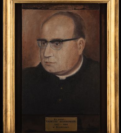 Zdjęcie nr 3: Portret uzyskał kształt stojącego prostokąta ujętego w złoconą, profilowaną ramę. Na ciemnym, brązowawym tle przedstawiono w popiersiu duchownego ukazanego w trzech czwartych w prawo. Jest to dojrzały mężczyzna w okularach o ciemnych oprawkach z owalną twarzą z wysokim łysiejącym czołem i z siwiejącymi, krótkimi włosami zaczesanymi do tyłu, odsłaniającymi lewe ucho. Jego twarz charakteryzują jasna karnacja, wydatny nos, wąskie usta i szeroko otwarte ciemne oczy. Ubrany jest w czarną sutannę z białą koloratką pod szyją. Pod portretem umieszczono tabliczkę z napisem "Ks(iądz) prałat / TADEUSZ BIEŃKOWSKI / 1963-1984 / w Brzeszczach". 