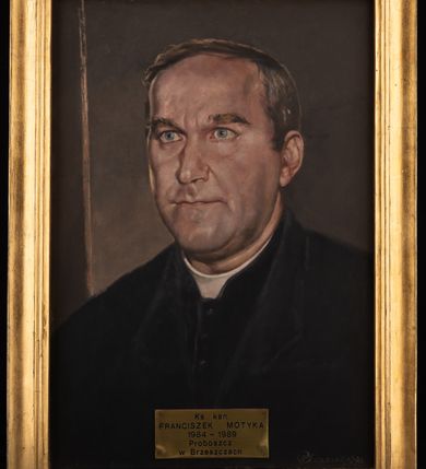 Zdjęcie nr 2: Portret uzyskał kształt stojącego prostokąta ujętego w złoconą, profilowaną ramę. Na ciemnym, brązowawym tle z zaznaczoną jasną krawędzią abstrakcyjnej przestrzeni, przedstawiono w popiersiu duchownego ukazanego w trzech czwartych w prawo. Jest to dojrzały mężczyzna z pociągłą twarzą z wysokim czołem z siwiejącymi, krótkimi włosami zaczesanymi w grzywkę z przedziałkiem, odsłaniającymi lewe ucho. Jego twarz charakteryzują jasna karnacja, wydatny nos, wąskie usta i szeroko otwarte niebieskie oczy oraz krzaczaste brwi. Ubrany jest w czarną sutannę z białą koloratką pod szyją oraz czarny płaszcz. Pod portretem umieszczono tabliczkę z napisem "Ks(siądz) kan(onik) / FRANCISZEK MOTYKA / 1984-1989 / Proboszcz / w Brzeszczach". 