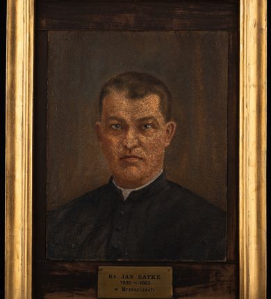 Zdjęcie nr 2: Portret uzyskał kształt stojącego prostokąta ujętego w złoconą, profilowaną ramę. Duchownego przedstawiono frontalnie w popiersiu na ciemnym tle. Jest to dojrzały mężczyzna z pociągłą twarzą z wysokim czołem z widocznymi nieznacznymi zakolami ciemnych, krótkich włosów odsłaniających oba ucha. Jego twarz charakteryzują jasna karnacja, mięsisty nos i wydatne usta  oraz niebieskie oczy o spojrzeniu skierowanym na widza. Ubrany jest w czarną sutannę z białą koloratką pod szyją. Pod portretem umieszczono tabliczkę z napisem "Ks(iądz) JAN SATKE / 1900-1903 / w Brzeszczach". 
