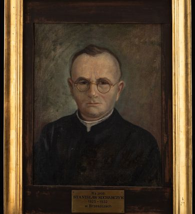 Zdjęcie nr 2: Portret uzyskał kształt stojącego prostokąta ujętego w złoconą, profilowaną ramę. Na ciemnym tle nieznacznie rozjaśnionym wokoło głowy przedstawiono w popiersiu duchownego nieznacznie ujętego w trzech czwartych w lewo. Jest to dojrzały mężczyzna w okrągłych okularach o złocistych oprawkach z pociągłą twarzą, bardzo wysokim czołem, z krótkimi, szpakowatymi włosami zaczesanymi do tyłu. Jego twarz charakteryzują jasna karnacja, wydatny nos, wąskie usta, ciemne oczy i krzaczaste brwi. Widoczne są także uszy. Ubrany jest w czarną sutannę z białą koloratką pod szyją. Pod portretem znajduje się tabliczka z napisem &quot;Ks(iądz) prob(oszcz) / STANISŁAW KUCHARCZYK / 1925-1932 / w Brzeszczach&quot;.  