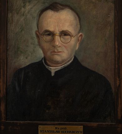 Zdjęcie nr 1: Portret uzyskał kształt stojącego prostokąta ujętego w złoconą, profilowaną ramę. Na ciemnym tle nieznacznie rozjaśnionym wokoło głowy przedstawiono w popiersiu duchownego nieznacznie ujętego w trzech czwartych w lewo. Jest to dojrzały mężczyzna w okrągłych okularach o złocistych oprawkach z pociągłą twarzą, bardzo wysokim czołem, z krótkimi, szpakowatymi włosami zaczesanymi do tyłu. Jego twarz charakteryzują jasna karnacja, wydatny nos, wąskie usta, ciemne oczy i krzaczaste brwi. Widoczne są także uszy. Ubrany jest w czarną sutannę z białą koloratką pod szyją. Pod portretem znajduje się tabliczka z napisem &quot;Ks(iądz) prob(oszcz) / STANISŁAW KUCHARCZYK / 1925-1932 / w Brzeszczach&quot;.  