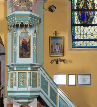 Zdjęcie nr 1: Przyścienna, wisząca ambona składa się z kosza i prowadzących do niego jednobiegowych schodów z balustradą, zaplecka oraz baldachimu. Wieloboczny kosz ustawiono na profilowanym, łamanym cokole. Poniżej widoczne jest podwieszenie utworzone ze zmniejszających się profilów układających się w spłaszczony stożek. Boki kosza artykułowano za pomocą diagonalnie ustawionych filarków na cokolikach podtrzymujących imposty z profilowanym i łamanym belkowaniem. Trzony filarków ozdobiono prostokątnymi płycinami z rozetą w środku. Płaszczyzny pomiędzy filarkami udekorowano prostokątnymi płycinami obwiedzionymi obramieniami z wolich oczu z geometryzowanym, płaskorzeźbionym ornamentem roślinno-kwiatowym w polu. Płyciny pomiędzy impostami przy górnych krawędziach ozdobiono ornamentem kostkowym. Zaplecek ambony ujęto dwoma pilastrami o płaskich głowicach zdobionych motywem stylizowanych liści.  Trzony pilastrów przedzielono na środku medalionem z rozetą i dwiema palmetami. Pomiędzy pilastrami umieszczono pole obrazowe w kształcie wydłużonego prostokąta o ściętych narożach w części górnej. W polu znajduje się obraz z całopostaciowym przedstawieniem Chrystusa jako Dobrego Pasterza. Ponad zapleckiem zawieszono wieloboczny baldachim utworzony z łamanego, profilowanego belkowania i podwieszonego lambrekinu, zwieńczony wielobocznymi spływami, na szczycie których  umieszczono na poduszce zamkniętą księgę, a za nią na obłokach w otoczeniu promieni dwie tablice Dekalogu. Strukturę architektoniczną ambony polichromowano w kolorze opalizującej zieleni, profile, ornamenty wyzłocona, a tablice Dekalogu i obłoki posrebrzono.     