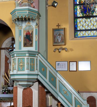 Zdjęcie nr 2: Przyścienna, wisząca ambona składa się z kosza i prowadzących do niego jednobiegowych schodów z balustradą, zaplecka oraz baldachimu. Wieloboczny kosz ustawiono na profilowanym, łamanym cokole. Poniżej widoczne jest podwieszenie utworzone ze zmniejszających się profilów układających się w spłaszczony stożek. Boki kosza artykułowano za pomocą diagonalnie ustawionych filarków na cokolikach podtrzymujących imposty z profilowanym i łamanym belkowaniem. Trzony filarków ozdobiono prostokątnymi płycinami z rozetą w środku. Płaszczyzny pomiędzy filarkami udekorowano prostokątnymi płycinami obwiedzionymi obramieniami z wolich oczu z geometryzowanym, płaskorzeźbionym ornamentem roślinno-kwiatowym w polu. Płyciny pomiędzy impostami przy górnych krawędziach ozdobiono ornamentem kostkowym. Zaplecek ambony ujęto dwoma pilastrami o płaskich głowicach zdobionych motywem stylizowanych liści.  Trzony pilastrów przedzielono na środku medalionem z rozetą i dwiema palmetami. Pomiędzy pilastrami umieszczono pole obrazowe w kształcie wydłużonego prostokąta o ściętych narożach w części górnej. W polu znajduje się obraz z całopostaciowym przedstawieniem Chrystusa jako Dobrego Pasterza. Ponad zapleckiem zawieszono wieloboczny baldachim utworzony z łamanego, profilowanego belkowania i podwieszonego lambrekinu, zwieńczony wielobocznymi spływami, na szczycie których  umieszczono na poduszce zamkniętą księgę, a za nią na obłokach w otoczeniu promieni dwie tablice Dekalogu. Strukturę architektoniczną ambony polichromowano w kolorze opalizującej zieleni, profile, ornamenty wyzłocona, a tablice Dekalogu i obłoki posrebrzono.     