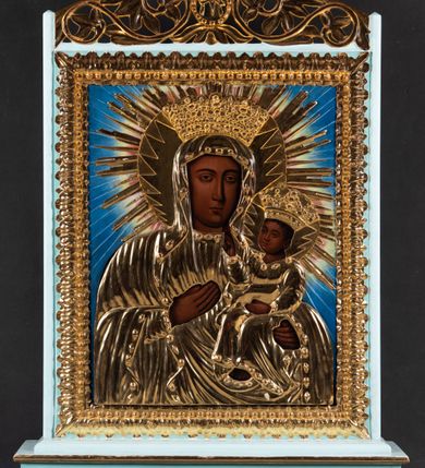 Zdjęcie nr 3: Feretron tworzy dwustronne pole obrazowe w kształcie stojącego prostokąta, zamknięte w ramę dekorowaną motywem palmetowym, ujęte z trzech stron fazowanymi listwami. Górną listwę udekorowano ażurowym, złoconym ornamentem utworzonym ze stylizowanej wici roślinnej flankującej owalny medalion z chrystogramem &quot;IHS&quot; na awersie i monogramem maryjnym na rewersie, zwieńczonym krzyżem. Całość przymocowana jest do prostopadłościennego, wydłużonego i profilowanego postumentu przeprutego po bokach otworami na drążki do noszenia. Dolną krawędź postumentu dekoruje płaskorzeźbiony ornament z motywem liścia palmetowego. Na awersie umieszczono obraz w kształcie stojącego prostokąta z przedstawieniem Najświętszego Serca Pana Jezusa. Zbawiciel został ukazany frontalnie w dwóch trzecich postaci. Jezus by móc ukazać swoje Boskie Serce rozsuwa na piersi obiema przebitymi rękami tunikę. Płonące serce ściśnięte korona cierniową otaczają złociste promienie. Z rany oraz u góry serca widać wydobywające się płomienie. Twarz Chrystusa o jasnej karnacji i rumianych policzkach jest pociągła. Otaczają je długie brązowe włosy spływające puklami na ramiona oraz brązowa, krótka broda. Charakteryzuje się ona wysokim czołem, długim, wąskim nosem i wydatnymi ustami. Chrystus szeroko otwartymi niebieskimi oczami wpatruje się w widza. Jego głowę otaczają złociste promienie. Ubrany jest w luźną, tunikę przewiązaną w pasie szarfą i narzucony na ramiona płaszcz, którego prawa poła przerzucona jest przez prawe przedramię. Zbawiciel został ukazany na jasnobrązowym tle. Partie szat przykrywa drewniana, złocona sukienka zdobiona w wypukłe kwiatuszki. Podobnie i namalowane promienie przykryto złoconymi, profilowanymi promieniami ostro zakończonymi w jaskółczy ogon. Na rewersie pola obrazowego feretronu umieszczono wizerunek Matki Boskiej Częstochowskiej. Madonna została ukazana frontalnie w półpostaci. Prawą ręką wskazuje na trzymane na lewym ramieniu Dzieciątko Jezus. Jej podłużną twarz o ciemnej karnacji charakteryzuje wydatny, prosty nos, migdałowe oczy, mięsiste usta oraz dwa równoległe cięcia widoczne na prawym policzku. Na głowie ma maforion lamowany taśmą z klejnotami, odsłaniający fragment sukni na piersi oraz prawy jej rękaw. Maria ma na głowie wysoką koronę o obłych sterczynach dekorowaną imitacją pereł oraz kamieni szlachetnych. Dzieciątko zostało ujęte w trzech czwartych z głową zwróconą w lewą stronę. Prawą rękę wznosi w geście błogosławieństwa, w lewej natomiast trzyma gruby kodeks Ewangelii. Mały Jezus ma owalną twarz o ciemnej karnacji. Widoczny jest wydatny nos i usta oraz szeroko otwarte oczy. Ma na sobie tunikę lamowaną taśmą z klejnotami. Głowę Chrystusa wieńczy wysoka korona, kształtem i dekoracją zbliżona do korony Madonny. Głowy postaci otaczają zębate nimby wpisane w okręgi, poszerzone o czerwonożółty otok ze złotymi promieniami widocznymi na jasnoniebieskim tle obrazu. Obie postacie przykrywają złocone sukienki wotywne odsłaniające jedynie partie twarzy, dłoni i stóp oraz korony i nimby z ostro zakończonymi promieniami.