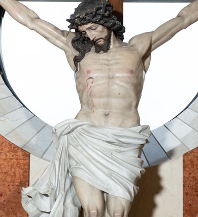 Zdjęcie nr 2: Na krzyżu z jasnego drewna zawieszono polichromowaną figurę martwego Chrystusa o szeroko rozpostartych ramionach. Ciało Zbawiciela do krzyża jest przymocowane za pomocą trzech gwoździ. On sam został ukazany jako martwy z zamkniętymi oczami i nieznacznie słaniającą się głową. Jego postać jest nieznacznie wysmuklona z naturalnie potraktowaną muskulaturą korpusu, zaznaczononą raną prawego boku. Nogi są lekko zwrółcone do osi środkowej, z założeniem prawej stopy na lewą. Twarz Zbawiciela jest mocno wychudła i pociągła. Charakteryzują ją zamknięte oczy, wydatny nos i usta. Otaczają ją brązowe włosy opadające puklami na kark oraz na prawe ramię oraz  brązowy zarost. Chrystus ma na głowie cierniową koronę, spod której na czoło spływają nieznaczne krople krwi. Biodra Zbawiciela przepasane są szarobiałym perizonium w formie zawoju z rozwianym festonem przy prawym boku. Tkaninę drapują ostre załamania. Ponad głową Chrystusa na pionowej belce krzyża zawieszono rozwiany titulus z napisem INRI przytwierdzone do krzyża wbitym na środku gwoździem. Całą figurę pokrywa naturalistyczna polichromia.