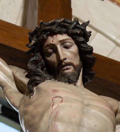Zdjęcie nr 4: Na krzyżu z jasnego drewna zawieszono polichromowaną figurę martwego Chrystusa o szeroko rozpostartych ramionach. Ciało Zbawiciela do krzyża jest przymocowane za pomocą trzech gwoździ. On sam został ukazany jako martwy z zamkniętymi oczami i nieznacznie słaniającą się głową. Jego postać jest nieznacznie wysmuklona z naturalnie potraktowaną muskulaturą korpusu, zaznaczononą raną prawego boku. Nogi są lekko zwrółcone do osi środkowej, z założeniem prawej stopy na lewą. Twarz Zbawiciela jest mocno wychudła i pociągła. Charakteryzują ją zamknięte oczy, wydatny nos i usta. Otaczają ją brązowe włosy opadające puklami na kark oraz na prawe ramię oraz  brązowy zarost. Chrystus ma na głowie cierniową koronę, spod której na czoło spływają nieznaczne krople krwi. Biodra Zbawiciela przepasane są szarobiałym perizonium w formie zawoju z rozwianym festonem przy prawym boku. Tkaninę drapują ostre załamania. Ponad głową Chrystusa na pionowej belce krzyża zawieszono rozwiany titulus z napisem INRI przytwierdzone do krzyża wbitym na środku gwoździem. Całą figurę pokrywa naturalistyczna polichromia.