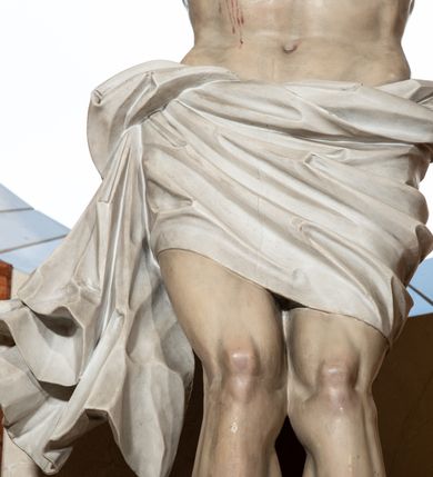 Zdjęcie nr 5: Na krzyżu z jasnego drewna zawieszono polichromowaną figurę martwego Chrystusa o szeroko rozpostartych ramionach. Ciało Zbawiciela do krzyża jest przymocowane za pomocą trzech gwoździ. On sam został ukazany jako martwy z zamkniętymi oczami i nieznacznie słaniającą się głową. Jego postać jest nieznacznie wysmuklona z naturalnie potraktowaną muskulaturą korpusu, zaznaczononą raną prawego boku. Nogi są lekko zwrółcone do osi środkowej, z założeniem prawej stopy na lewą. Twarz Zbawiciela jest mocno wychudła i pociągła. Charakteryzują ją zamknięte oczy, wydatny nos i usta. Otaczają ją brązowe włosy opadające puklami na kark oraz na prawe ramię oraz  brązowy zarost. Chrystus ma na głowie cierniową koronę, spod której na czoło spływają nieznaczne krople krwi. Biodra Zbawiciela przepasane są szarobiałym perizonium w formie zawoju z rozwianym festonem przy prawym boku. Tkaninę drapują ostre załamania. Ponad głową Chrystusa na pionowej belce krzyża zawieszono rozwiany titulus z napisem INRI przytwierdzone do krzyża wbitym na środku gwoździem. Całą figurę pokrywa naturalistyczna polichromia.