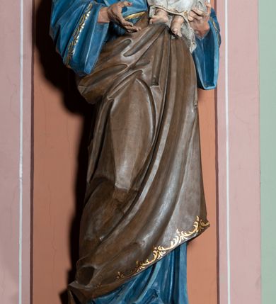 Zdjęcie nr 3: Pełnoplastyczna, rzeźba przedstawia w całej postaci ujętego w kontrapoście św. Józefa jako starszego mężczyznę stojącego na niskim czworobocznym postumencie o ściętych narożach. Józef na lewym przedramieniu trzyma Dzieciątko Jezus, wyciągając również przed siebie prawą rękę. Jego głowa ujęta została w trzech czwartych w prawo. Święty ma pociągłą twarz o ostrych rysach, jasnej karnacji, zapadłych policzkach z wydatnym nosem i ustami oraz spojrzeniem półprzymkniętych oczu skierowanych na Dzieciątko. Jego głowę otaczają ostro modelowane ciemne włosy i broda. Józef ubrany jest w niebieską tunikę lamowaną pod szyją i przy rękawach złotym ornamentem oraz w brązowy płaszcz przerzucony przez lewe ramię, którego prawa poła podwinięta jest na lewym boku. Mały Jezus został ukazany w trzech czwartych w lewo z rozłożonym obiema rękami i podkurczonymi nogami. Jego głowa widoczna z profilu skierowana jest w stronę św. Józefa. Jezus ma pociągłą twarz o jasnej karnacji i rumianych policzkach, wydatny nos i usta. Jego głowę otaczają krótkie, kędzierzawe jasne włosy. Ubrany jest w jasnoszarą tunikę lamowano pod szyją, na rękawach i na spodzie złotym obszyciem, która odsłania jego bose stopy. Na piersi Dzieciątka widoczne jest      płonące Boskie Serce z koroną cierniową, otoczone złocistymi promieniami. 