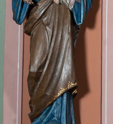 Zdjęcie nr 2: Pełnoplastyczna, rzeźba przedstawia w całej postaci ujętego w kontrapoście św. Józefa jako starszego mężczyznę stojącego na niskim czworobocznym postumencie o ściętych narożach. Józef na lewym przedramieniu trzyma Dzieciątko Jezus, wyciągając również przed siebie prawą rękę. Jego głowa ujęta została w trzech czwartych w prawo. Święty ma pociągłą twarz o ostrych rysach, jasnej karnacji, zapadłych policzkach z wydatnym nosem i ustami oraz spojrzeniem półprzymkniętych oczu skierowanych na Dzieciątko. Jego głowę otaczają ostro modelowane ciemne włosy i broda. Józef ubrany jest w niebieską tunikę lamowaną pod szyją i przy rękawach złotym ornamentem oraz w brązowy płaszcz przerzucony przez lewe ramię, którego prawa poła podwinięta jest na lewym boku. Mały Jezus został ukazany w trzech czwartych w lewo z rozłożonym obiema rękami i podkurczonymi nogami. Jego głowa widoczna z profilu skierowana jest w stronę św. Józefa. Jezus ma pociągłą twarz o jasnej karnacji i rumianych policzkach, wydatny nos i usta. Jego głowę otaczają krótkie, kędzierzawe jasne włosy. Ubrany jest w jasnoszarą tunikę lamowano pod szyją, na rękawach i na spodzie złotym obszyciem, która odsłania jego bose stopy. Na piersi Dzieciątka widoczne jest      płonące Boskie Serce z koroną cierniową, otoczone złocistymi promieniami. 