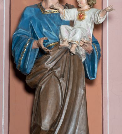 Zdjęcie nr 4: Pełnoplastyczna, rzeźba przedstawia w całej postaci ujętego w kontrapoście św. Józefa jako starszego mężczyznę stojącego na niskim czworobocznym postumencie o ściętych narożach. Józef na lewym przedramieniu trzyma Dzieciątko Jezus, wyciągając również przed siebie prawą rękę. Jego głowa ujęta została w trzech czwartych w prawo. Święty ma pociągłą twarz o ostrych rysach, jasnej karnacji, zapadłych policzkach z wydatnym nosem i ustami oraz spojrzeniem półprzymkniętych oczu skierowanych na Dzieciątko. Jego głowę otaczają ostro modelowane ciemne włosy i broda. Józef ubrany jest w niebieską tunikę lamowaną pod szyją i przy rękawach złotym ornamentem oraz w brązowy płaszcz przerzucony przez lewe ramię, którego prawa poła podwinięta jest na lewym boku. Mały Jezus został ukazany w trzech czwartych w lewo z rozłożonym obiema rękami i podkurczonymi nogami. Jego głowa widoczna z profilu skierowana jest w stronę św. Józefa. Jezus ma pociągłą twarz o jasnej karnacji i rumianych policzkach, wydatny nos i usta. Jego głowę otaczają krótkie, kędzierzawe jasne włosy. Ubrany jest w jasnoszarą tunikę lamowano pod szyją, na rękawach i na spodzie złotym obszyciem, która odsłania jego bose stopy. Na piersi Dzieciątka widoczne jest      płonące Boskie Serce z koroną cierniową, otoczone złocistymi promieniami. 