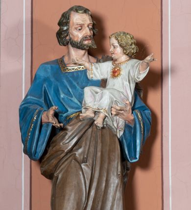 Zdjęcie nr 1: Pełnoplastyczna, rzeźba przedstawia w całej postaci ujętego w kontrapoście św. Józefa jako starszego mężczyznę stojącego na niskim czworobocznym postumencie o ściętych narożach. Józef na lewym przedramieniu trzyma Dzieciątko Jezus, wyciągając również przed siebie prawą rękę. Jego głowa ujęta została w trzech czwartych w prawo. Święty ma pociągłą twarz o ostrych rysach, jasnej karnacji, zapadłych policzkach z wydatnym nosem i ustami oraz spojrzeniem półprzymkniętych oczu skierowanych na Dzieciątko. Jego głowę otaczają ostro modelowane ciemne włosy i broda. Józef ubrany jest w niebieską tunikę lamowaną pod szyją i przy rękawach złotym ornamentem oraz w brązowy płaszcz przerzucony przez lewe ramię, którego prawa poła podwinięta jest na lewym boku. Mały Jezus został ukazany w trzech czwartych w lewo z rozłożonym obiema rękami i podkurczonymi nogami. Jego głowa widoczna z profilu skierowana jest w stronę św. Józefa. Jezus ma pociągłą twarz o jasnej karnacji i rumianych policzkach, wydatny nos i usta. Jego głowę otaczają krótkie, kędzierzawe jasne włosy. Ubrany jest w jasnoszarą tunikę lamowano pod szyją, na rękawach i na spodzie złotym obszyciem, która odsłania jego bose stopy. Na piersi Dzieciątka widoczne jest      płonące Boskie Serce z koroną cierniową, otoczone złocistymi promieniami. 
