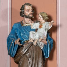 Zdjęcie nr 1: Pełnoplastyczna, rzeźba przedstawia w całej postaci ujętego w kontrapoście św. Józefa jako starszego mężczyznę stojącego na niskim czworobocznym postumencie o ściętych narożach. Józef na lewym przedramieniu trzyma Dzieciątko Jezus, wyciągając również przed siebie prawą rękę. Jego głowa ujęta została w trzech czwartych w prawo. Święty ma pociągłą twarz o ostrych rysach, jasnej karnacji, zapadłych policzkach z wydatnym nosem i ustami oraz spojrzeniem półprzymkniętych oczu skierowanych na Dzieciątko. Jego głowę otaczają ostro modelowane ciemne włosy i broda. Józef ubrany jest w niebieską tunikę lamowaną pod szyją i przy rękawach złotym ornamentem oraz w brązowy płaszcz przerzucony przez lewe ramię, którego prawa poła podwinięta jest na lewym boku. Mały Jezus został ukazany w trzech czwartych w lewo z rozłożonym obiema rękami i podkurczonymi nogami. Jego głowa widoczna z profilu skierowana jest w stronę św. Józefa. Jezus ma pociągłą twarz o jasnej karnacji i rumianych policzkach, wydatny nos i usta. Jego głowę otaczają krótkie, kędzierzawe jasne włosy. Ubrany jest w jasnoszarą tunikę lamowano pod szyją, na rękawach i na spodzie złotym obszyciem, która odsłania jego bose stopy. Na piersi Dzieciątka widoczne jest      płonące Boskie Serce z koroną cierniową, otoczone złocistymi promieniami. 