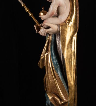 Zdjęcie nr 7: Rzeźba pełnoplastyczna, przyścienna. Przedstawienie Matki Boskiej z Dzieciątkiem ukazanej frontalnie, w całej postaci, w postawie stojącej, z ugiętymi w łokciach rękami; w prawej dłoni trzyma przed sobą berło, prawą obejmuje Dzieciątko. Twarz owalna, o jasnej karnacji, drobnych rysach, z wysokim czołem, okolona włosami spływającymi na ramiona i plecy. Na głowie złota otwarta korona (wtórna). Ubrana w długą suknię z długimi rękawami, zmarszczoną pionowymi równoległymi fałdami, przewiązaną w pasie. Na ramiona nałożony złoty płaszcz z błękitną podszewką, okrywający całą postać i odwijający się pod prawą ręką i przy podstawie okazałymi fałdami muszlowymi. Nagie Dzieciątko o pulchnym ciele siedzi na lewym przedramieniu Matki. Kulista głowa na krótkiej szyi okolona kędzierzawymi włosami. Prawą dłonią błogosławi, w lewej trzyma kulę. 