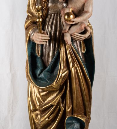 Zdjęcie nr 2: Rzeźba pełnoplastyczna, przyścienna. Przedstawienie Matki Boskiej z Dzieciątkiem ukazanej frontalnie, w całej postaci, w postawie stojącej, z ugiętymi w łokciach rękami; w prawej dłoni trzyma przed sobą berło, prawą obejmuje Dzieciątko. Twarz owalna, o jasnej karnacji, drobnych rysach, z wysokim czołem, okolona włosami spływającymi na ramiona i plecy. Na głowie złota otwarta korona (wtórna). Ubrana w długą suknię z długimi rękawami, zmarszczoną pionowymi równoległymi fałdami, przewiązaną w pasie. Na ramiona nałożony złoty płaszcz z błękitną podszewką, okrywający całą postać i odwijający się pod prawą ręką i przy podstawie okazałymi fałdami muszlowymi. Nagie Dzieciątko o pulchnym ciele siedzi na lewym przedramieniu Matki. Kulista głowa na krótkiej szyi okolona kędzierzawymi włosami. Prawą dłonią błogosławi, w lewej trzyma kulę. 