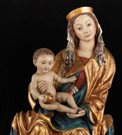Zdjęcie nr 1: Rzeźba pełnoplastyczna, przyścienna, z tyłu wydrążona. Całopostaciowe przedstawienie tronującej Matki Boskiej z Dzieciątkiem. Maria, ukazana frontalnie, jako młoda kobieta, siedzi z rozstawionymi kolanami, z łukowato przegiętym w lewo tułowiem i głową lekko przechyloną w prawo. Ugiętymi w łokciach rękami podtrzymuje przed sobą Dzieciątko. Twarz Marii owalna, pełna, o jasnej karnacji, z szeroko rozstawionymi oczami, regularnymi łukami brwiowymi i refleksem uśmiechu na ustach. Głowę okrywa srebrzona chusta, opadająca na ramiona dwiema kaskadami rurkowatych fałdów, spod której widać krótkie brązowe włosy. Na głowie niski złoty diadem. Maria ubrana w długą srebrną, przewiązaną w pasie suknię, spod fałdów której widać czubki srebrnych butów. Na ramiona ma narzucony złocony płaszcz spięty na piersiach i okrywający kaskadami dekoracyjnych fałdów biodra i nogi Marii oraz ławę, na której siedzi. Nagie Dzieciątko o dużej głowie i wątłym ciele siedzi bokiem na kolanach Marii, twarz zwracając ku widzowi. Lewą ręką dotyka piersi Matki, w prawej trzyma błękitną kulę. 