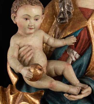 Zdjęcie nr 12: Rzeźba pełnoplastyczna, przyścienna, z tyłu wydrążona. Całopostaciowe przedstawienie tronującej Matki Boskiej z Dzieciątkiem. Maria, ukazana frontalnie, jako młoda kobieta, siedzi z rozstawionymi kolanami, z łukowato przegiętym w lewo tułowiem i głową lekko przechyloną w prawo. Ugiętymi w łokciach rękami podtrzymuje przed sobą Dzieciątko. Twarz Marii owalna, pełna, o jasnej karnacji, z szeroko rozstawionymi oczami, regularnymi łukami brwiowymi i refleksem uśmiechu na ustach. Głowę okrywa srebrzona chusta, opadająca na ramiona dwiema kaskadami rurkowatych fałdów, spod której widać krótkie brązowe włosy. Na głowie niski złoty diadem. Maria ubrana w długą srebrną, przewiązaną w pasie suknię, spod fałdów której widać czubki srebrnych butów. Na ramiona ma narzucony złocony płaszcz spięty na piersiach i okrywający kaskadami dekoracyjnych fałdów biodra i nogi Marii oraz ławę, na której siedzi. Nagie Dzieciątko o dużej głowie i wątłym ciele siedzi bokiem na kolanach Marii, twarz zwracając ku widzowi. Lewą ręką dotyka piersi Matki, w prawej trzyma błękitną kulę. 