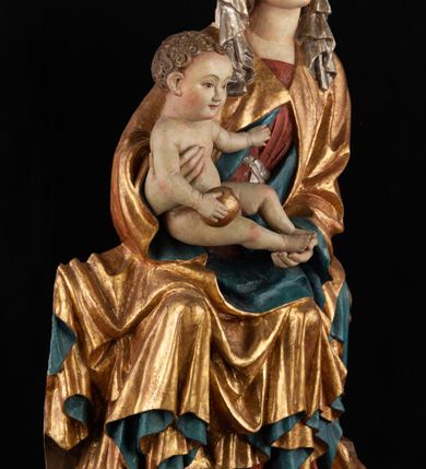 Zdjęcie nr 2: Rzeźba pełnoplastyczna, przyścienna, z tyłu wydrążona. Całopostaciowe przedstawienie tronującej Matki Boskiej z Dzieciątkiem. Maria, ukazana frontalnie, jako młoda kobieta, siedzi z rozstawionymi kolanami, z łukowato przegiętym w lewo tułowiem i głową lekko przechyloną w prawo. Ugiętymi w łokciach rękami podtrzymuje przed sobą Dzieciątko. Twarz Marii owalna, pełna, o jasnej karnacji, z szeroko rozstawionymi oczami, regularnymi łukami brwiowymi i refleksem uśmiechu na ustach. Głowę okrywa srebrzona chusta, opadająca na ramiona dwiema kaskadami rurkowatych fałdów, spod której widać krótkie brązowe włosy. Na głowie niski złoty diadem. Maria ubrana w długą srebrną, przewiązaną w pasie suknię, spod fałdów której widać czubki srebrnych butów. Na ramiona ma narzucony złocony płaszcz spięty na piersiach i okrywający kaskadami dekoracyjnych fałdów biodra i nogi Marii oraz ławę, na której siedzi. Nagie Dzieciątko o dużej głowie i wątłym ciele siedzi bokiem na kolanach Marii, twarz zwracając ku widzowi. Lewą ręką dotyka piersi Matki, w prawej trzyma błękitną kulę. 