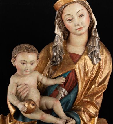Zdjęcie nr 7: Rzeźba pełnoplastyczna, przyścienna, z tyłu wydrążona. Całopostaciowe przedstawienie tronującej Matki Boskiej z Dzieciątkiem. Maria, ukazana frontalnie, jako młoda kobieta, siedzi z rozstawionymi kolanami, z łukowato przegiętym w lewo tułowiem i głową lekko przechyloną w prawo. Ugiętymi w łokciach rękami podtrzymuje przed sobą Dzieciątko. Twarz Marii owalna, pełna, o jasnej karnacji, z szeroko rozstawionymi oczami, regularnymi łukami brwiowymi i refleksem uśmiechu na ustach. Głowę okrywa srebrzona chusta, opadająca na ramiona dwiema kaskadami rurkowatych fałdów, spod której widać krótkie brązowe włosy. Na głowie niski złoty diadem. Maria ubrana w długą srebrną, przewiązaną w pasie suknię, spod fałdów której widać czubki srebrnych butów. Na ramiona ma narzucony złocony płaszcz spięty na piersiach i okrywający kaskadami dekoracyjnych fałdów biodra i nogi Marii oraz ławę, na której siedzi. Nagie Dzieciątko o dużej głowie i wątłym ciele siedzi bokiem na kolanach Marii, twarz zwracając ku widzowi. Lewą ręką dotyka piersi Matki, w prawej trzyma błękitną kulę. 