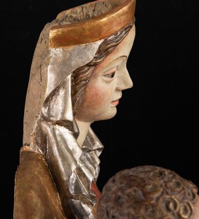 Zdjęcie nr 11: Rzeźba pełnoplastyczna, przyścienna, z tyłu wydrążona. Całopostaciowe przedstawienie tronującej Matki Boskiej z Dzieciątkiem. Maria, ukazana frontalnie, jako młoda kobieta, siedzi z rozstawionymi kolanami, z łukowato przegiętym w lewo tułowiem i głową lekko przechyloną w prawo. Ugiętymi w łokciach rękami podtrzymuje przed sobą Dzieciątko. Twarz Marii owalna, pełna, o jasnej karnacji, z szeroko rozstawionymi oczami, regularnymi łukami brwiowymi i refleksem uśmiechu na ustach. Głowę okrywa srebrzona chusta, opadająca na ramiona dwiema kaskadami rurkowatych fałdów, spod której widać krótkie brązowe włosy. Na głowie niski złoty diadem. Maria ubrana w długą srebrną, przewiązaną w pasie suknię, spod fałdów której widać czubki srebrnych butów. Na ramiona ma narzucony złocony płaszcz spięty na piersiach i okrywający kaskadami dekoracyjnych fałdów biodra i nogi Marii oraz ławę, na której siedzi. Nagie Dzieciątko o dużej głowie i wątłym ciele siedzi bokiem na kolanach Marii, twarz zwracając ku widzowi. Lewą ręką dotyka piersi Matki, w prawej trzyma błękitną kulę. 