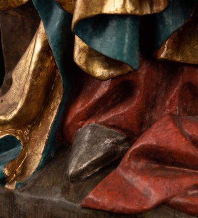 Zdjęcie nr 14: Rzeźba pełnoplastyczna, przyścienna, z tyłu wydrążona. Całopostaciowe przedstawienie tronującej Matki Boskiej z Dzieciątkiem. Maria, ukazana frontalnie, jako młoda kobieta, siedzi z rozstawionymi kolanami, z łukowato przegiętym w lewo tułowiem i głową lekko przechyloną w prawo. Ugiętymi w łokciach rękami podtrzymuje przed sobą Dzieciątko. Twarz Marii owalna, pełna, o jasnej karnacji, z szeroko rozstawionymi oczami, regularnymi łukami brwiowymi i refleksem uśmiechu na ustach. Głowę okrywa srebrzona chusta, opadająca na ramiona dwiema kaskadami rurkowatych fałdów, spod której widać krótkie brązowe włosy. Na głowie niski złoty diadem. Maria ubrana w długą srebrną, przewiązaną w pasie suknię, spod fałdów której widać czubki srebrnych butów. Na ramiona ma narzucony złocony płaszcz spięty na piersiach i okrywający kaskadami dekoracyjnych fałdów biodra i nogi Marii oraz ławę, na której siedzi. Nagie Dzieciątko o dużej głowie i wątłym ciele siedzi bokiem na kolanach Marii, twarz zwracając ku widzowi. Lewą ręką dotyka piersi Matki, w prawej trzyma błękitną kulę. 