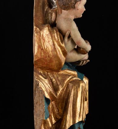 Zdjęcie nr 16: Rzeźba pełnoplastyczna, przyścienna, z tyłu wydrążona. Całopostaciowe przedstawienie tronującej Matki Boskiej z Dzieciątkiem. Maria, ukazana frontalnie, jako młoda kobieta, siedzi z rozstawionymi kolanami, z łukowato przegiętym w lewo tułowiem i głową lekko przechyloną w prawo. Ugiętymi w łokciach rękami podtrzymuje przed sobą Dzieciątko. Twarz Marii owalna, pełna, o jasnej karnacji, z szeroko rozstawionymi oczami, regularnymi łukami brwiowymi i refleksem uśmiechu na ustach. Głowę okrywa srebrzona chusta, opadająca na ramiona dwiema kaskadami rurkowatych fałdów, spod której widać krótkie brązowe włosy. Na głowie niski złoty diadem. Maria ubrana w długą srebrną, przewiązaną w pasie suknię, spod fałdów której widać czubki srebrnych butów. Na ramiona ma narzucony złocony płaszcz spięty na piersiach i okrywający kaskadami dekoracyjnych fałdów biodra i nogi Marii oraz ławę, na której siedzi. Nagie Dzieciątko o dużej głowie i wątłym ciele siedzi bokiem na kolanach Marii, twarz zwracając ku widzowi. Lewą ręką dotyka piersi Matki, w prawej trzyma błękitną kulę. 