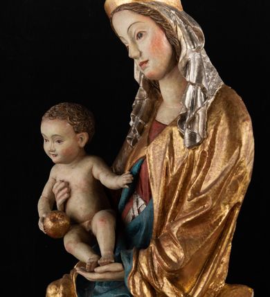 Zdjęcie nr 4: Rzeźba pełnoplastyczna, przyścienna, z tyłu wydrążona. Całopostaciowe przedstawienie tronującej Matki Boskiej z Dzieciątkiem. Maria, ukazana frontalnie, jako młoda kobieta, siedzi z rozstawionymi kolanami, z łukowato przegiętym w lewo tułowiem i głową lekko przechyloną w prawo. Ugiętymi w łokciach rękami podtrzymuje przed sobą Dzieciątko. Twarz Marii owalna, pełna, o jasnej karnacji, z szeroko rozstawionymi oczami, regularnymi łukami brwiowymi i refleksem uśmiechu na ustach. Głowę okrywa srebrzona chusta, opadająca na ramiona dwiema kaskadami rurkowatych fałdów, spod której widać krótkie brązowe włosy. Na głowie niski złoty diadem. Maria ubrana w długą srebrną, przewiązaną w pasie suknię, spod fałdów której widać czubki srebrnych butów. Na ramiona ma narzucony złocony płaszcz spięty na piersiach i okrywający kaskadami dekoracyjnych fałdów biodra i nogi Marii oraz ławę, na której siedzi. Nagie Dzieciątko o dużej głowie i wątłym ciele siedzi bokiem na kolanach Marii, twarz zwracając ku widzowi. Lewą ręką dotyka piersi Matki, w prawej trzyma błękitną kulę. 