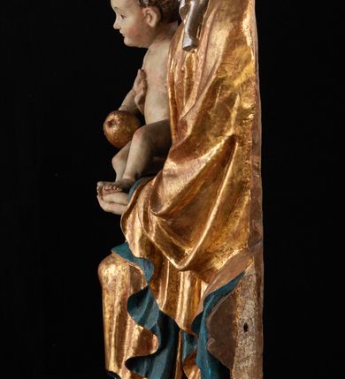 Zdjęcie nr 17: Rzeźba pełnoplastyczna, przyścienna, z tyłu wydrążona. Całopostaciowe przedstawienie tronującej Matki Boskiej z Dzieciątkiem. Maria, ukazana frontalnie, jako młoda kobieta, siedzi z rozstawionymi kolanami, z łukowato przegiętym w lewo tułowiem i głową lekko przechyloną w prawo. Ugiętymi w łokciach rękami podtrzymuje przed sobą Dzieciątko. Twarz Marii owalna, pełna, o jasnej karnacji, z szeroko rozstawionymi oczami, regularnymi łukami brwiowymi i refleksem uśmiechu na ustach. Głowę okrywa srebrzona chusta, opadająca na ramiona dwiema kaskadami rurkowatych fałdów, spod której widać krótkie brązowe włosy. Na głowie niski złoty diadem. Maria ubrana w długą srebrną, przewiązaną w pasie suknię, spod fałdów której widać czubki srebrnych butów. Na ramiona ma narzucony złocony płaszcz spięty na piersiach i okrywający kaskadami dekoracyjnych fałdów biodra i nogi Marii oraz ławę, na której siedzi. Nagie Dzieciątko o dużej głowie i wątłym ciele siedzi bokiem na kolanach Marii, twarz zwracając ku widzowi. Lewą ręką dotyka piersi Matki, w prawej trzyma błękitną kulę. 