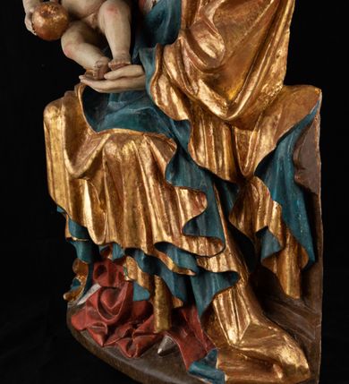 Zdjęcie nr 18: Rzeźba pełnoplastyczna, przyścienna, z tyłu wydrążona. Całopostaciowe przedstawienie tronującej Matki Boskiej z Dzieciątkiem. Maria, ukazana frontalnie, jako młoda kobieta, siedzi z rozstawionymi kolanami, z łukowato przegiętym w lewo tułowiem i głową lekko przechyloną w prawo. Ugiętymi w łokciach rękami podtrzymuje przed sobą Dzieciątko. Twarz Marii owalna, pełna, o jasnej karnacji, z szeroko rozstawionymi oczami, regularnymi łukami brwiowymi i refleksem uśmiechu na ustach. Głowę okrywa srebrzona chusta, opadająca na ramiona dwiema kaskadami rurkowatych fałdów, spod której widać krótkie brązowe włosy. Na głowie niski złoty diadem. Maria ubrana w długą srebrną, przewiązaną w pasie suknię, spod fałdów której widać czubki srebrnych butów. Na ramiona ma narzucony złocony płaszcz spięty na piersiach i okrywający kaskadami dekoracyjnych fałdów biodra i nogi Marii oraz ławę, na której siedzi. Nagie Dzieciątko o dużej głowie i wątłym ciele siedzi bokiem na kolanach Marii, twarz zwracając ku widzowi. Lewą ręką dotyka piersi Matki, w prawej trzyma błękitną kulę. 