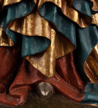 Zdjęcie nr 19: Rzeźba pełnoplastyczna, przyścienna, z tyłu wydrążona. Całopostaciowe przedstawienie tronującej Matki Boskiej z Dzieciątkiem. Maria, ukazana frontalnie, jako młoda kobieta, siedzi z rozstawionymi kolanami, z łukowato przegiętym w lewo tułowiem i głową lekko przechyloną w prawo. Ugiętymi w łokciach rękami podtrzymuje przed sobą Dzieciątko. Twarz Marii owalna, pełna, o jasnej karnacji, z szeroko rozstawionymi oczami, regularnymi łukami brwiowymi i refleksem uśmiechu na ustach. Głowę okrywa srebrzona chusta, opadająca na ramiona dwiema kaskadami rurkowatych fałdów, spod której widać krótkie brązowe włosy. Na głowie niski złoty diadem. Maria ubrana w długą srebrną, przewiązaną w pasie suknię, spod fałdów której widać czubki srebrnych butów. Na ramiona ma narzucony złocony płaszcz spięty na piersiach i okrywający kaskadami dekoracyjnych fałdów biodra i nogi Marii oraz ławę, na której siedzi. Nagie Dzieciątko o dużej głowie i wątłym ciele siedzi bokiem na kolanach Marii, twarz zwracając ku widzowi. Lewą ręką dotyka piersi Matki, w prawej trzyma błękitną kulę. 