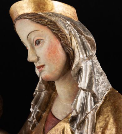 Zdjęcie nr 10: Rzeźba pełnoplastyczna, przyścienna, z tyłu wydrążona. Całopostaciowe przedstawienie tronującej Matki Boskiej z Dzieciątkiem. Maria, ukazana frontalnie, jako młoda kobieta, siedzi z rozstawionymi kolanami, z łukowato przegiętym w lewo tułowiem i głową lekko przechyloną w prawo. Ugiętymi w łokciach rękami podtrzymuje przed sobą Dzieciątko. Twarz Marii owalna, pełna, o jasnej karnacji, z szeroko rozstawionymi oczami, regularnymi łukami brwiowymi i refleksem uśmiechu na ustach. Głowę okrywa srebrzona chusta, opadająca na ramiona dwiema kaskadami rurkowatych fałdów, spod której widać krótkie brązowe włosy. Na głowie niski złoty diadem. Maria ubrana w długą srebrną, przewiązaną w pasie suknię, spod fałdów której widać czubki srebrnych butów. Na ramiona ma narzucony złocony płaszcz spięty na piersiach i okrywający kaskadami dekoracyjnych fałdów biodra i nogi Marii oraz ławę, na której siedzi. Nagie Dzieciątko o dużej głowie i wątłym ciele siedzi bokiem na kolanach Marii, twarz zwracając ku widzowi. Lewą ręką dotyka piersi Matki, w prawej trzyma błękitną kulę. 