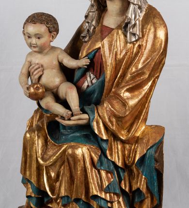 Zdjęcie nr 5: Rzeźba pełnoplastyczna, przyścienna, z tyłu wydrążona. Całopostaciowe przedstawienie tronującej Matki Boskiej z Dzieciątkiem. Maria, ukazana frontalnie, jako młoda kobieta, siedzi z rozstawionymi kolanami, z łukowato przegiętym w lewo tułowiem i głową lekko przechyloną w prawo. Ugiętymi w łokciach rękami podtrzymuje przed sobą Dzieciątko. Twarz Marii owalna, pełna, o jasnej karnacji, z szeroko rozstawionymi oczami, regularnymi łukami brwiowymi i refleksem uśmiechu na ustach. Głowę okrywa srebrzona chusta, opadająca na ramiona dwiema kaskadami rurkowatych fałdów, spod której widać krótkie brązowe włosy. Na głowie niski złoty diadem. Maria ubrana w długą srebrną, przewiązaną w pasie suknię, spod fałdów której widać czubki srebrnych butów. Na ramiona ma narzucony złocony płaszcz spięty na piersiach i okrywający kaskadami dekoracyjnych fałdów biodra i nogi Marii oraz ławę, na której siedzi. Nagie Dzieciątko o dużej głowie i wątłym ciele siedzi bokiem na kolanach Marii, twarz zwracając ku widzowi. Lewą ręką dotyka piersi Matki, w prawej trzyma błękitną kulę. 