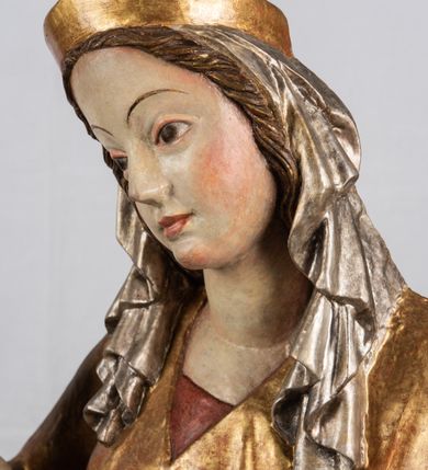 Zdjęcie nr 8: Rzeźba pełnoplastyczna, przyścienna, z tyłu wydrążona. Całopostaciowe przedstawienie tronującej Matki Boskiej z Dzieciątkiem. Maria, ukazana frontalnie, jako młoda kobieta, siedzi z rozstawionymi kolanami, z łukowato przegiętym w lewo tułowiem i głową lekko przechyloną w prawo. Ugiętymi w łokciach rękami podtrzymuje przed sobą Dzieciątko. Twarz Marii owalna, pełna, o jasnej karnacji, z szeroko rozstawionymi oczami, regularnymi łukami brwiowymi i refleksem uśmiechu na ustach. Głowę okrywa srebrzona chusta, opadająca na ramiona dwiema kaskadami rurkowatych fałdów, spod której widać krótkie brązowe włosy. Na głowie niski złoty diadem. Maria ubrana w długą srebrną, przewiązaną w pasie suknię, spod fałdów której widać czubki srebrnych butów. Na ramiona ma narzucony złocony płaszcz spięty na piersiach i okrywający kaskadami dekoracyjnych fałdów biodra i nogi Marii oraz ławę, na której siedzi. Nagie Dzieciątko o dużej głowie i wątłym ciele siedzi bokiem na kolanach Marii, twarz zwracając ku widzowi. Lewą ręką dotyka piersi Matki, w prawej trzyma błękitną kulę. 