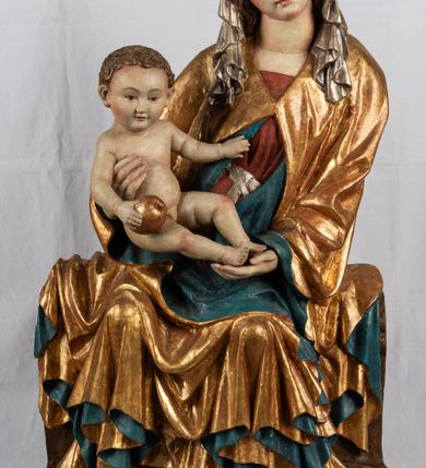 Zdjęcie nr 6: Rzeźba pełnoplastyczna, przyścienna, z tyłu wydrążona. Całopostaciowe przedstawienie tronującej Matki Boskiej z Dzieciątkiem. Maria, ukazana frontalnie, jako młoda kobieta, siedzi z rozstawionymi kolanami, z łukowato przegiętym w lewo tułowiem i głową lekko przechyloną w prawo. Ugiętymi w łokciach rękami podtrzymuje przed sobą Dzieciątko. Twarz Marii owalna, pełna, o jasnej karnacji, z szeroko rozstawionymi oczami, regularnymi łukami brwiowymi i refleksem uśmiechu na ustach. Głowę okrywa srebrzona chusta, opadająca na ramiona dwiema kaskadami rurkowatych fałdów, spod której widać krótkie brązowe włosy. Na głowie niski złoty diadem. Maria ubrana w długą srebrną, przewiązaną w pasie suknię, spod fałdów której widać czubki srebrnych butów. Na ramiona ma narzucony złocony płaszcz spięty na piersiach i okrywający kaskadami dekoracyjnych fałdów biodra i nogi Marii oraz ławę, na której siedzi. Nagie Dzieciątko o dużej głowie i wątłym ciele siedzi bokiem na kolanach Marii, twarz zwracając ku widzowi. Lewą ręką dotyka piersi Matki, w prawej trzyma błękitną kulę. 