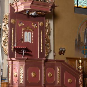 Zdjęcie nr 1: Ambona przyścienna, wisząca, z wejściem schodami od strony prezbiterium. Kosz na planie ośmioboku ze ściankami wydzielonymi pilastrami z prostokątnymi płycinami na trzonach. Pola ścianek wypełniają płyciny w formie stojącego prostokąta o ściętych narożach. Ujęty dołem wybrzuszonym architrawem parapet jest w formie niepełnego belkowania z gzymsem wyłamującym się nad pilastrami. U spodu kosza podwieszenie ujęte czterema wolutami, zakończone dekoracyjną szyszką w formie kiści winogrona. Schody są jednobiegowe z balustradą w formie ścianki wypełnionej romboidalną płyciną o ściętych narożach, ujętej na końcu kolumną o lekko wybrzuszonym trzonie, zwieńczoną niewielkim gazonem. Zaplecek wypełnia płycina w kształcie stojącego prostokąta. Baldachim na rzucie czworoboku o ściętych narożach, który u podstawy obiega wyłamujące się na narożach belkowanie, wieńczy forma czterech spływów wolutowych zakomponowanych naprzemiennie z trzema ażurowymi kartuszami w formie racaille&#039;ów. Strukturę kosza zdobią motywy rozet w płycinach ścianek i gałązek z rozetkami w płycinach pilastrów. Trzon kolumny balustrady oplata wić winnej latorośli. Płycinę zaplecka wypełnia polichromowany motyw tablic z przykazaniami Bożymi ujętych płaskorzeźbionym motywem liści akantu z parą kampanuli, w narożach jest para uskrzydlonych główek anielskich. Po bokach znajduje się para rozet i podwieszeń w formie ulistnionych gałązek z małymi rozetkami. Zaplecek ujmuje para ażurowych, ornamentalnych uszaków. Podniebie baldachimu wypełnia płaskorzeźbiony motyw gołębicy Ducha Świętego na tle promienistej glorii. Szczyt baldachimu jest w formie figury baranka paschalnego stojącego na zamkniętej księdze z siedmioma pieczęciami na podstawie z obłoków i ujętego półkolistą glorią promienistą. Struktura ambony utrzymana jest w odcieniach brązu, detal architektoniczny i ornamentyka są złocone i srebrzone.   