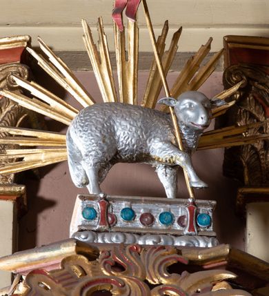 Zdjęcie nr 3: Ambona przyścienna, wisząca, z wejściem schodami od strony prezbiterium. Kosz na planie ośmioboku ze ściankami wydzielonymi pilastrami z prostokątnymi płycinami na trzonach. Pola ścianek wypełniają płyciny w formie stojącego prostokąta o ściętych narożach. Ujęty dołem wybrzuszonym architrawem parapet jest w formie niepełnego belkowania z gzymsem wyłamującym się nad pilastrami. U spodu kosza podwieszenie ujęte czterema wolutami, zakończone dekoracyjną szyszką w formie kiści winogrona. Schody są jednobiegowe z balustradą w formie ścianki wypełnionej romboidalną płyciną o ściętych narożach, ujętej na końcu kolumną o lekko wybrzuszonym trzonie, zwieńczoną niewielkim gazonem. Zaplecek wypełnia płycina w kształcie stojącego prostokąta. Baldachim na rzucie czworoboku o ściętych narożach, który u podstawy obiega wyłamujące się na narożach belkowanie, wieńczy forma czterech spływów wolutowych zakomponowanych naprzemiennie z trzema ażurowymi kartuszami w formie racaille'ów. Strukturę kosza zdobią motywy rozet w płycinach ścianek i gałązek z rozetkami w płycinach pilastrów. Trzon kolumny balustrady oplata wić winnej latorośli. Płycinę zaplecka wypełnia polichromowany motyw tablic z przykazaniami Bożymi ujętych płaskorzeźbionym motywem liści akantu z parą kampanuli, w narożach jest para uskrzydlonych główek anielskich. Po bokach znajduje się para rozet i podwieszeń w formie ulistnionych gałązek z małymi rozetkami. Zaplecek ujmuje para ażurowych, ornamentalnych uszaków. Podniebie baldachimu wypełnia płaskorzeźbiony motyw gołębicy Ducha Świętego na tle promienistej glorii. Szczyt baldachimu jest w formie figury baranka paschalnego stojącego na zamkniętej księdze z siedmioma pieczęciami na podstawie z obłoków i ujętego półkolistą glorią promienistą. Struktura ambony utrzymana jest w odcieniach brązu, detal architektoniczny i ornamentyka są złocone i srebrzone.   