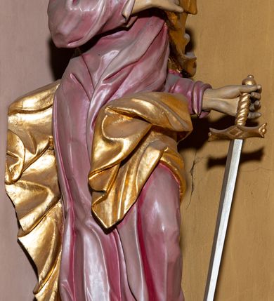 Zdjęcie nr 6: Rzeźba całopostaciowa przedstawia stojącego w wydatnym kontrapoście, na niewysokim postumencie św. Pawła zwróconego na wprost, z głową skierowaną w trzech czwartych w prawo. Twarz świętego charakteryzuje śniada karnacja, wyraziste rysy oraz długi, brązowy zarost. Półdługie, kręcone włosy opadają na kark mężczyzny. Święty ma na sobie długą, jasnoliliową suknię. Połę narzuconego na lewe ramię, rozwianego, złoconego płaszcza osłaniającego lewe biodro, podtrzymuje na wysokości klatki piersiowej prawą ręką. W lewej, zgiętej w łokciu i uniesionej na wysokości talii podtrzymuje miecz o złoconej rękojeści i srebrzonym ostrzu skierowanym w dół. 